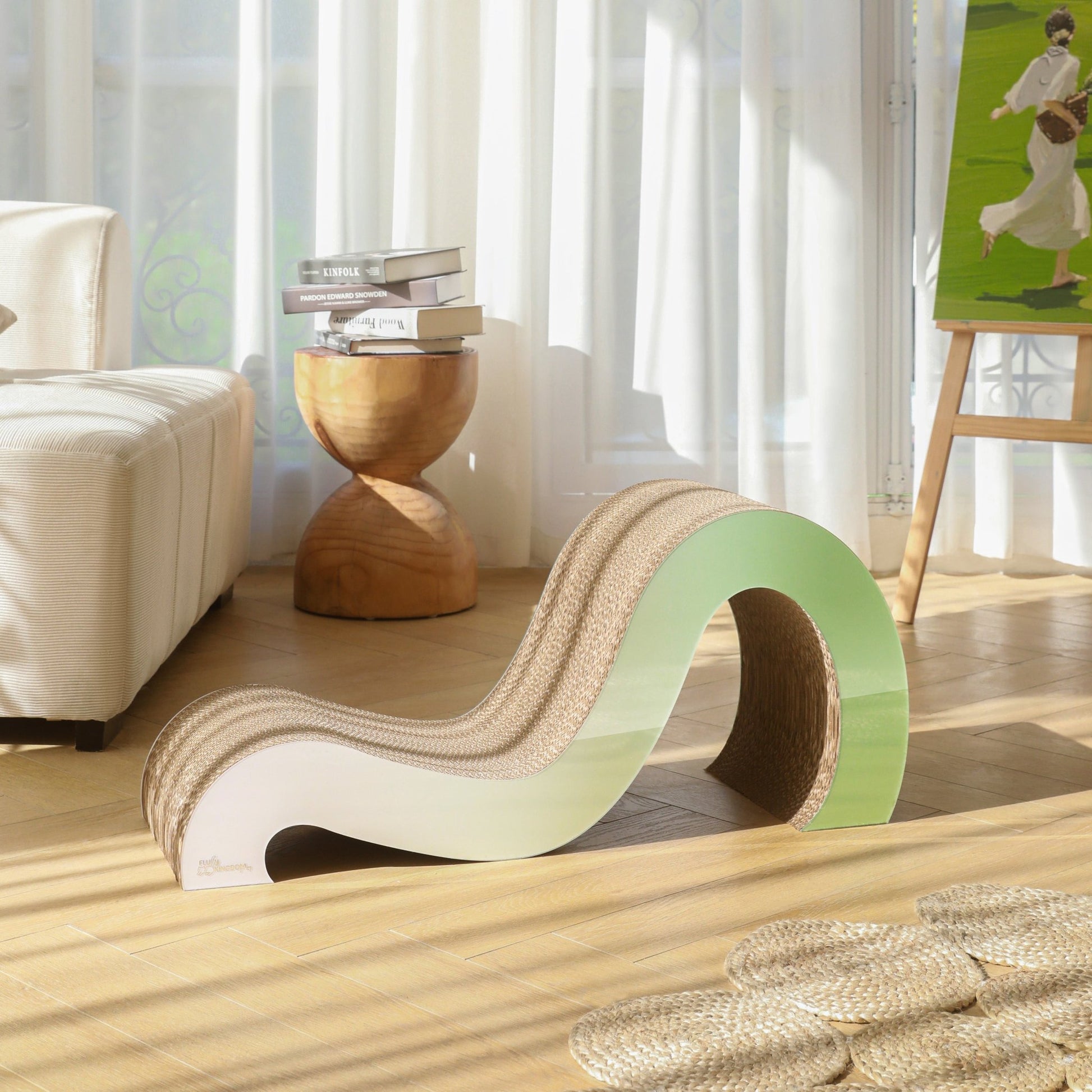 The Mint Mint - Modern Cat Scratcher & Stretching Board, crafted from premium cardboard in a wave shape, rests on a light wooden floor near a stack of books and a stool in a sunlit room with white curtains, ensuring healthy claws for your feline friend.