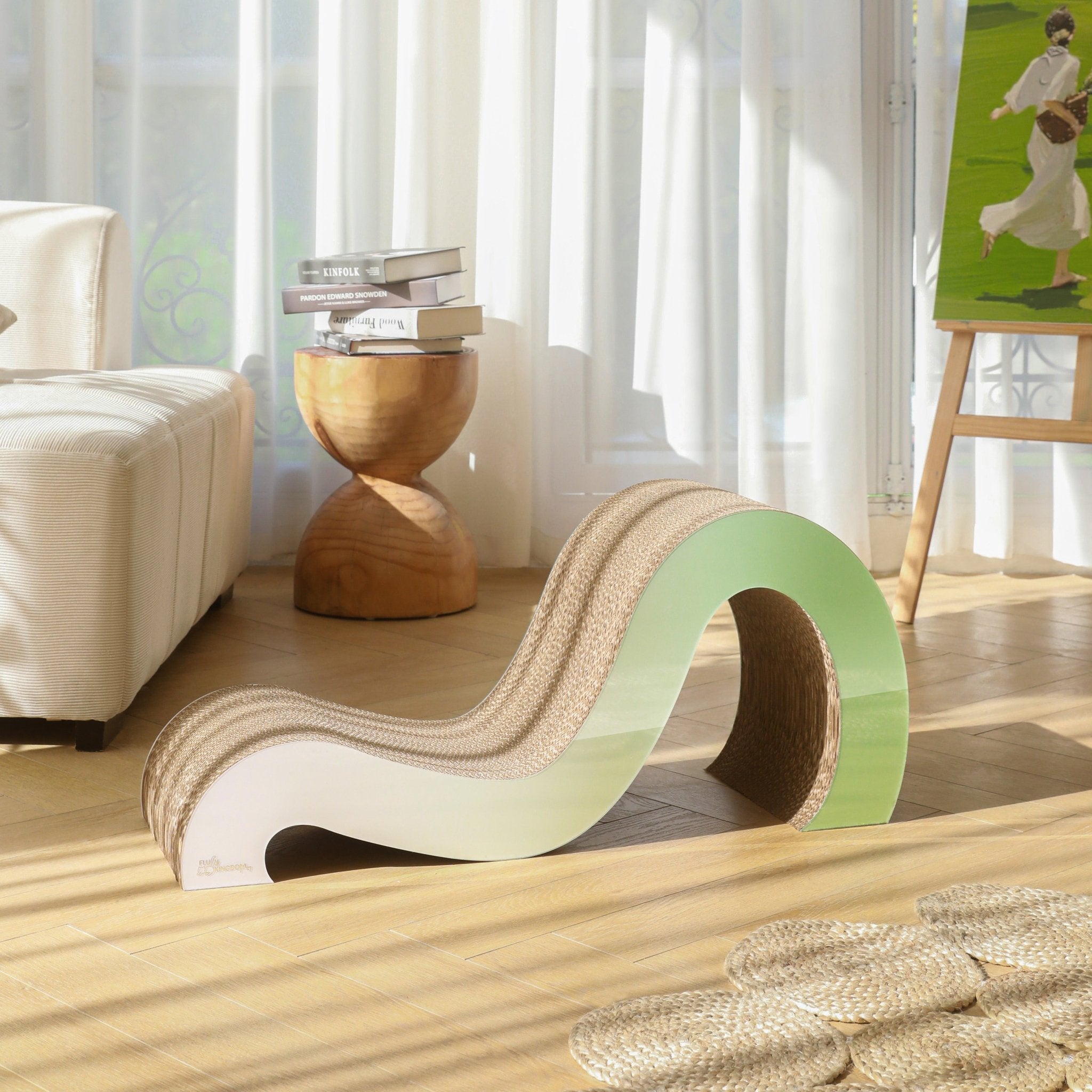 The Mint Mint - Modern Cat Scratcher & Stretching Board, crafted from premium cardboard in a wave shape, rests on a light wooden floor near a stack of books and a stool in a sunlit room with white curtains, ensuring healthy claws for your feline friend.