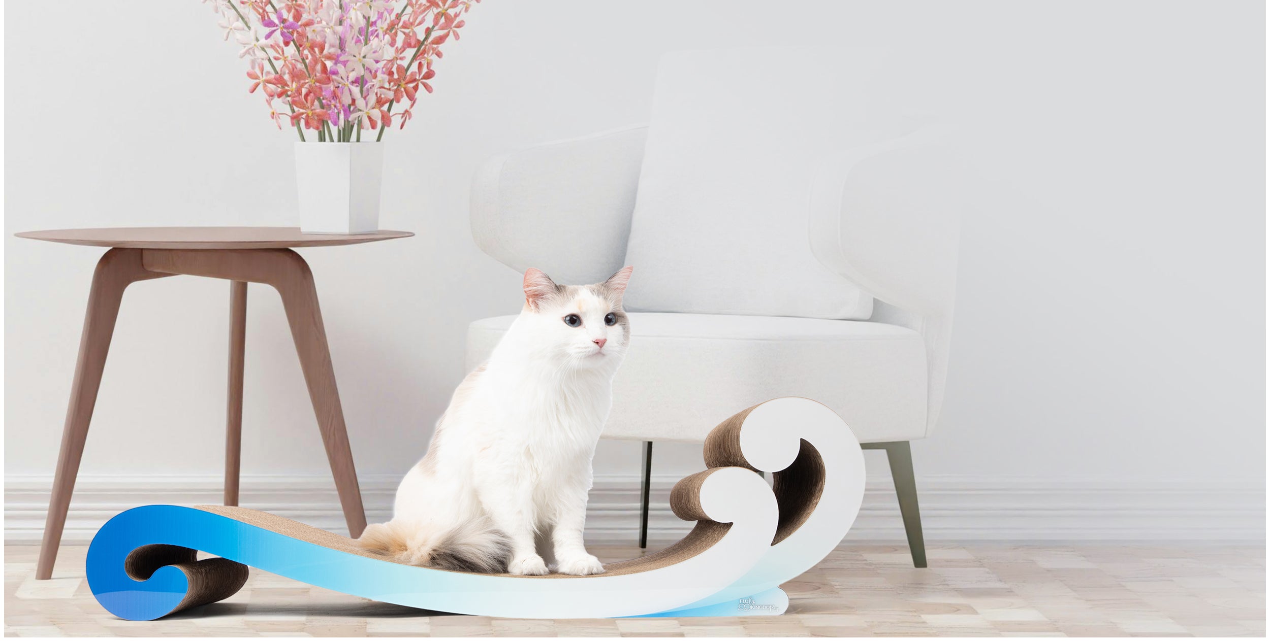 A white cat sits on a uniquely curved scratching board in a modern living room with a white armchair, side table, and a vase of pink flowers.