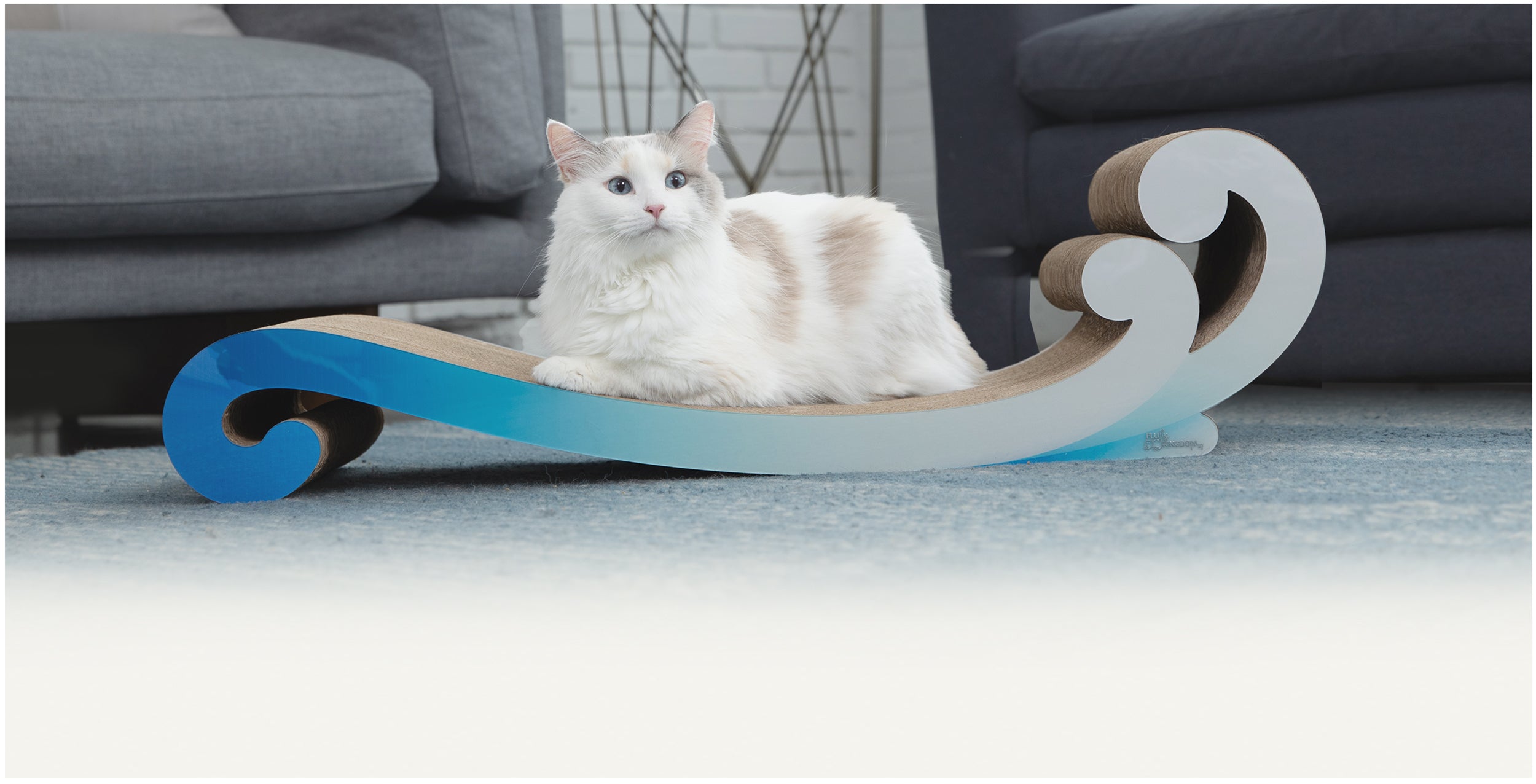 A white and gray cat lounges on a blue and white wave-shaped cat scratcher in a living room with gray furniture.