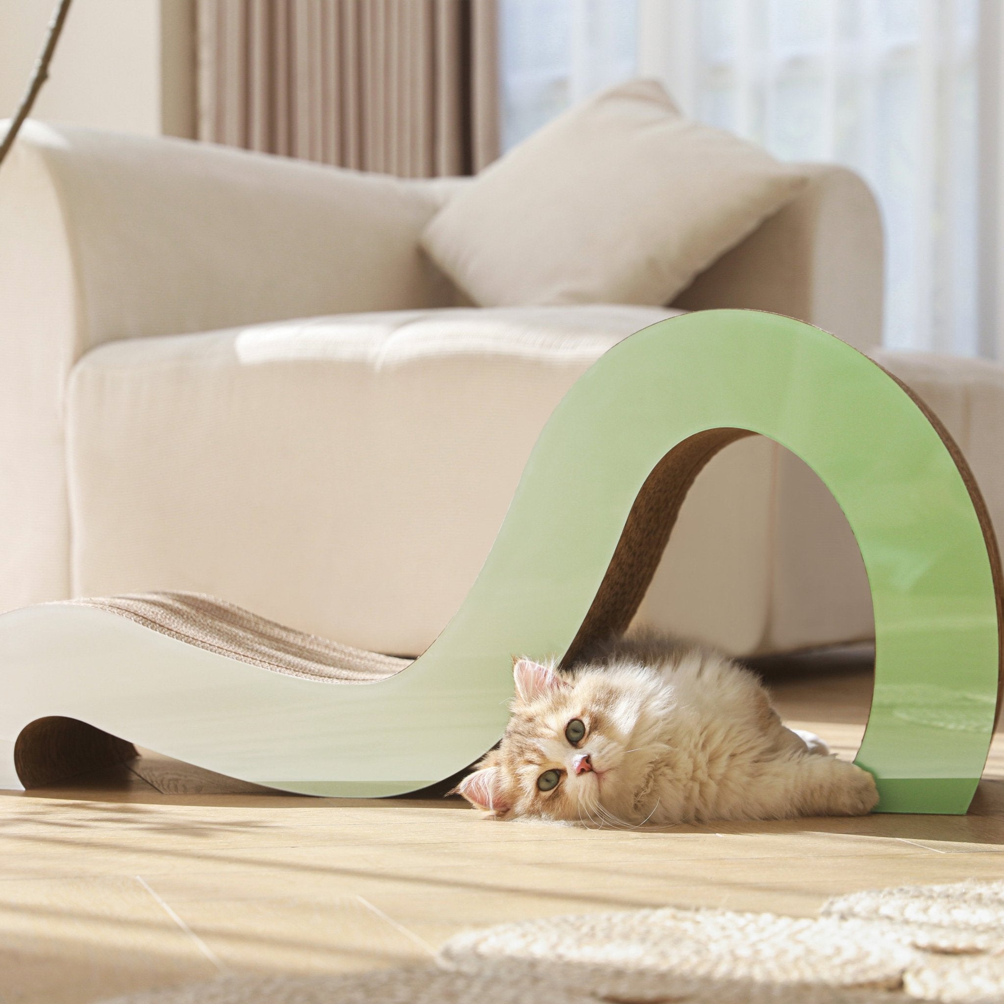 A fluffy cat lies on its side under the Mint Mint - Modern Cat Scratcher & Stretching Board, made of premium cardboard, in a sunlit, beige living room.